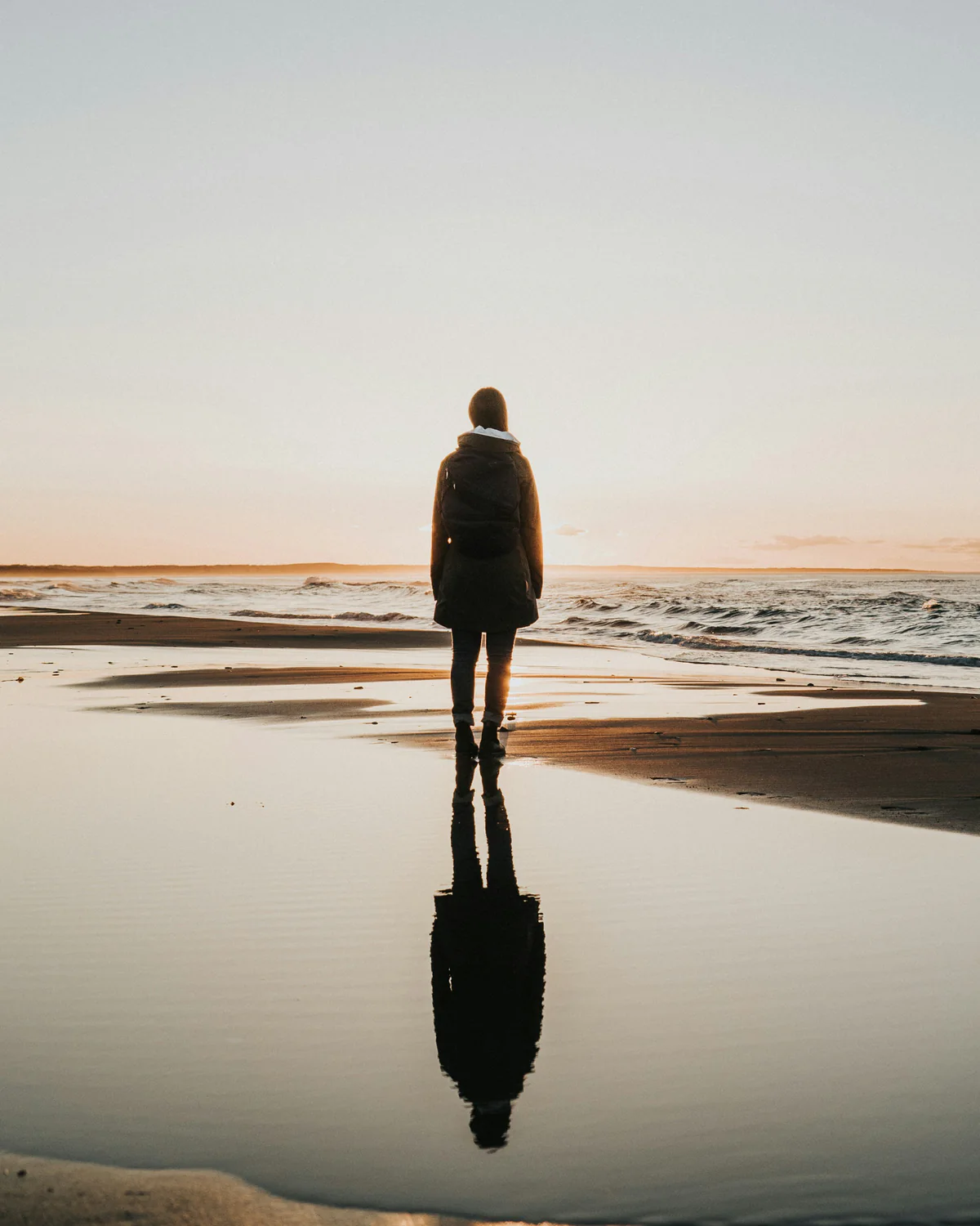 A person standing quietly at the shoreline, reflected in the wet sand